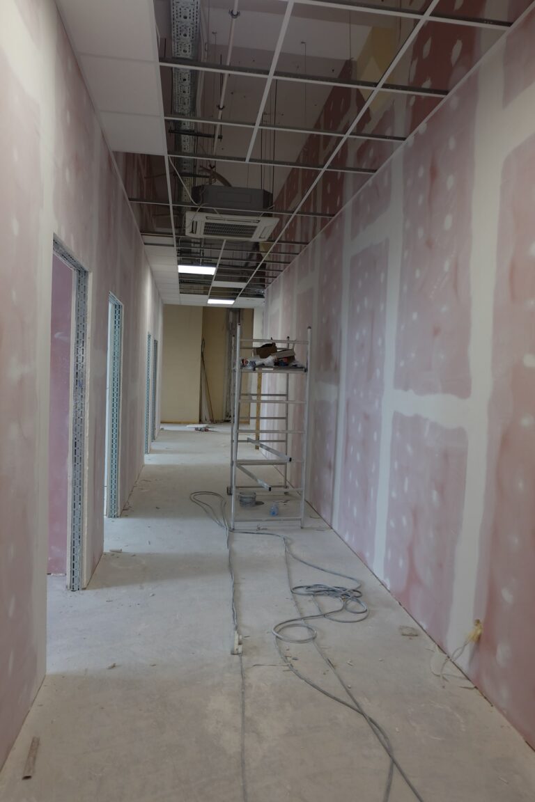 Na zdjęciu widać korytarz w czasie remontu. ściany są w gładzi. Na środku rozstawione jest rusztowanie.