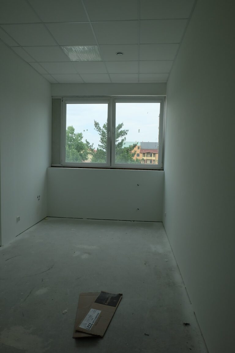 Na zdjęciu jest pokój z białymi ścianami i oknami wzdłuż jednej z nich. Pokój jest w remoncie.
