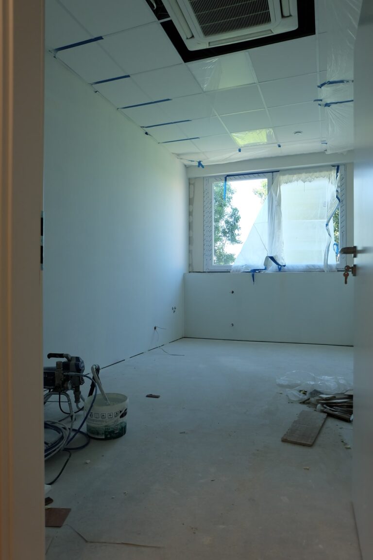 Na zdjęciu jest pokój z białymi ścianami i oknami wzdłuż jednej z nich. Pokój jest w remoncie. Na podłodze leżą narzędzia