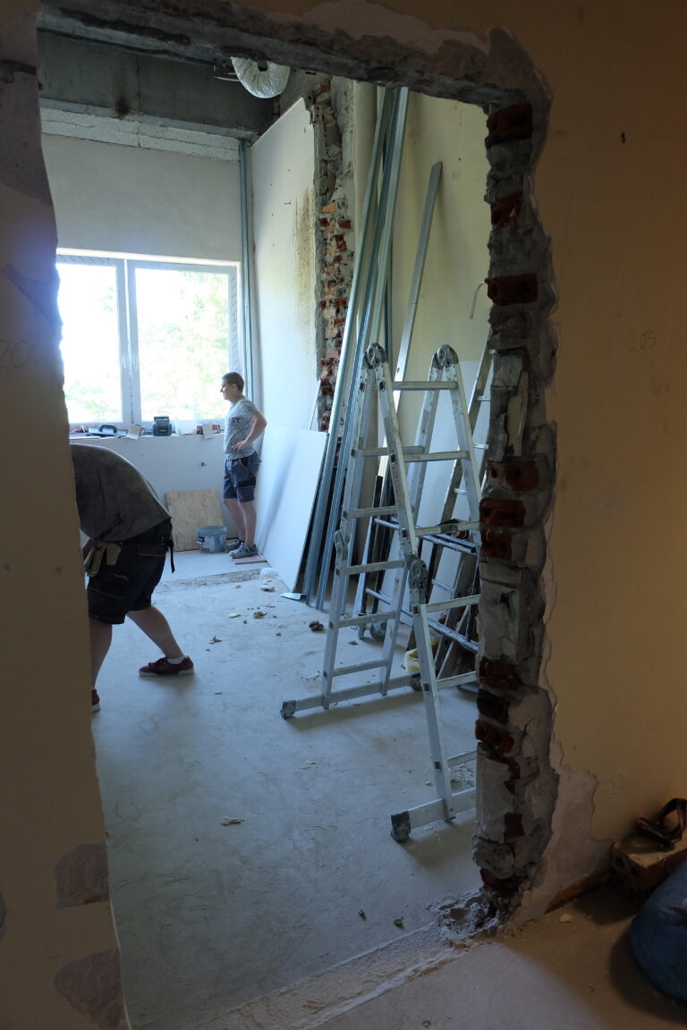 Na zadęciu widać prace remontowe, wejście do jednego z pomieszczeń oraz budowlańców. W pomieszczeniu znajduje się rozłożona srebrna drabina.