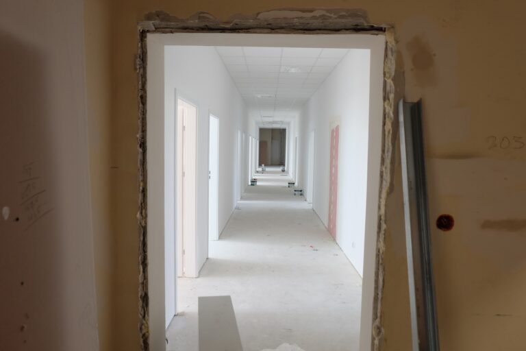 Na zdjęciu widać biały korytarz, który jest w remoncie. Wzdłuż ścian po obu stronach znajdują się drzwi gabinetów. Na podłodze rozstawione są wiaderka z farbą.
