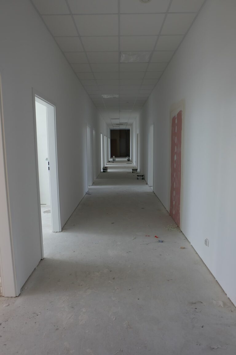Na zdjęciu widać biały korytarz, który jest w remoncie. Wzdłuż ścian po obu stronach znajdują się drzwi gabinetów. Na podłodze rozstawione są wiaderka z farbą.