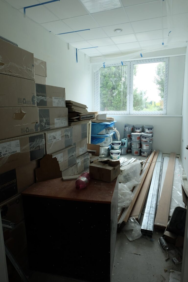 Na zdjęciu widać pokój z białymi ścianami i oknami na jednej ze ścian. W pokoju znajdują się kartony, wiaderka z farbami oraz inne narzędzia i materiały budowlane.