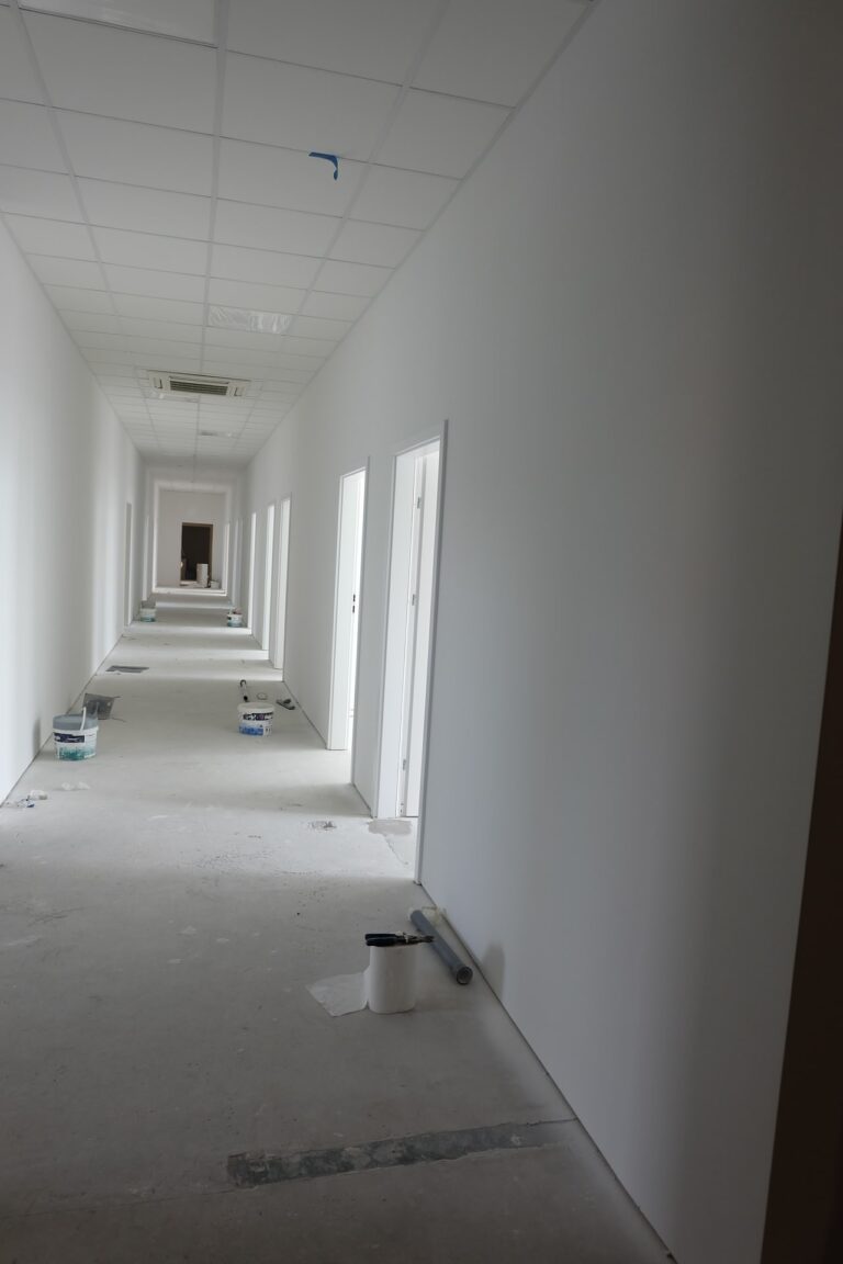 Na zdjęciu widać biały korytarz, który jest w remoncie. Wzdłuż ściany po prawej stronie znajdują się drzwi gabinetów. Na podłodze rozstawione są wiaderka z farbą.