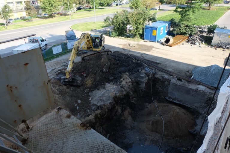 Zdjęcie przedstawia plac przed budynkiem. Prowadzone są prace budowlane, na środku zdjęcia jest wykopana dziura. Po jej lewej stronie stoi żółta koparka.