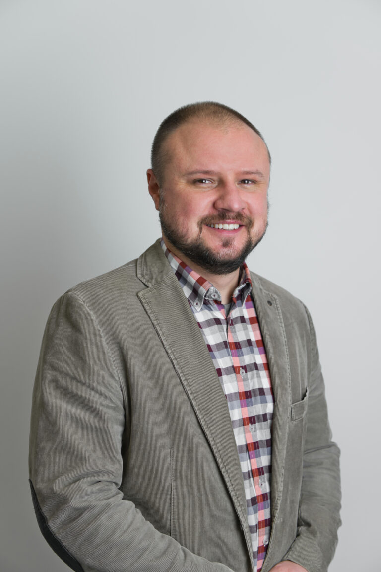 Zdjęcie przedstawia uśmiechnietego pana Witolda Wnukiewicza Mężczcyzna znajduje się na szarym tle. Ubrany jest w koszulę w kratke oraz szarą marynarkę