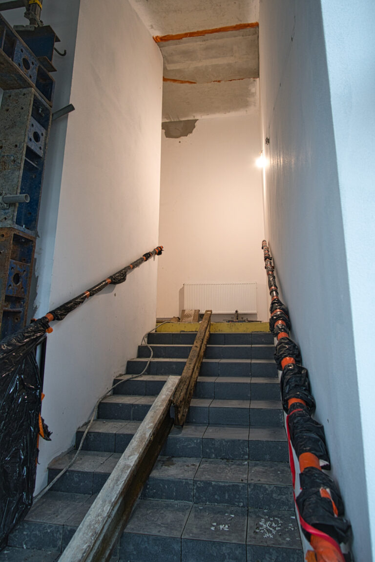 na zdjęciu widać klatkę schodową w czasie remontu na środku schodów są położone belki. na klatcce schodowej jest włączone światło