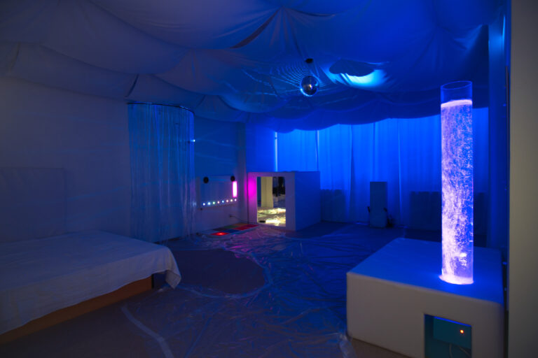 Zdjęcie przedstawia salę doświadczenia świata. Znajduje się tam łóżko wodne, lampę wodną i domek z lustrami Na podłodze jest rozłożona folia