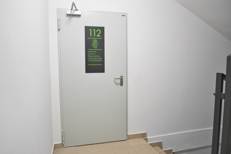 Na zdjęciu są szare duże drzwi z tabliczką z zielonym numerem 112 oraz logiem centrum