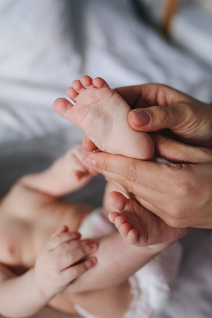 Na obrazie widać małe stopy dziecka, które są dotykane przez dłonie osoby dorosłej. dziecko jest widoczne od ramion w dół i jest ubrane w pampersa.