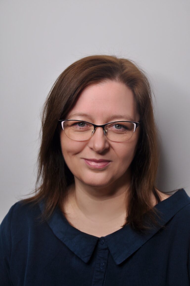 Zdjęcie przedstawia portret Pani Agnieszki Stemblewskiej na szarym tle. Ma ona okulary i brązowe włosy do ramion. Ubrana jest w granatowa koszulę z kołnierzem.